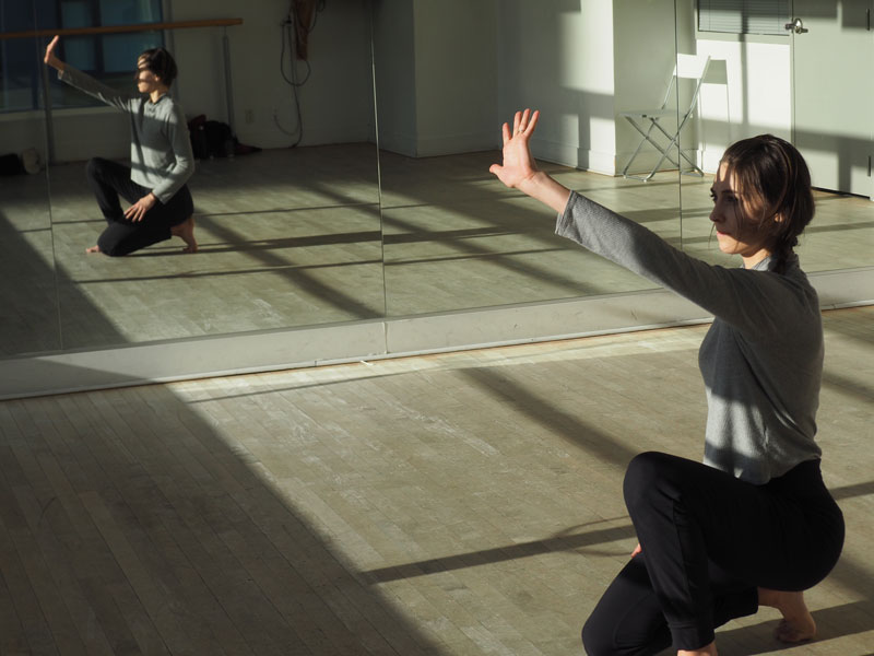 image of Rachel in a dance studio. There is sun coming in the window and Rachel has their hand in front of their face.