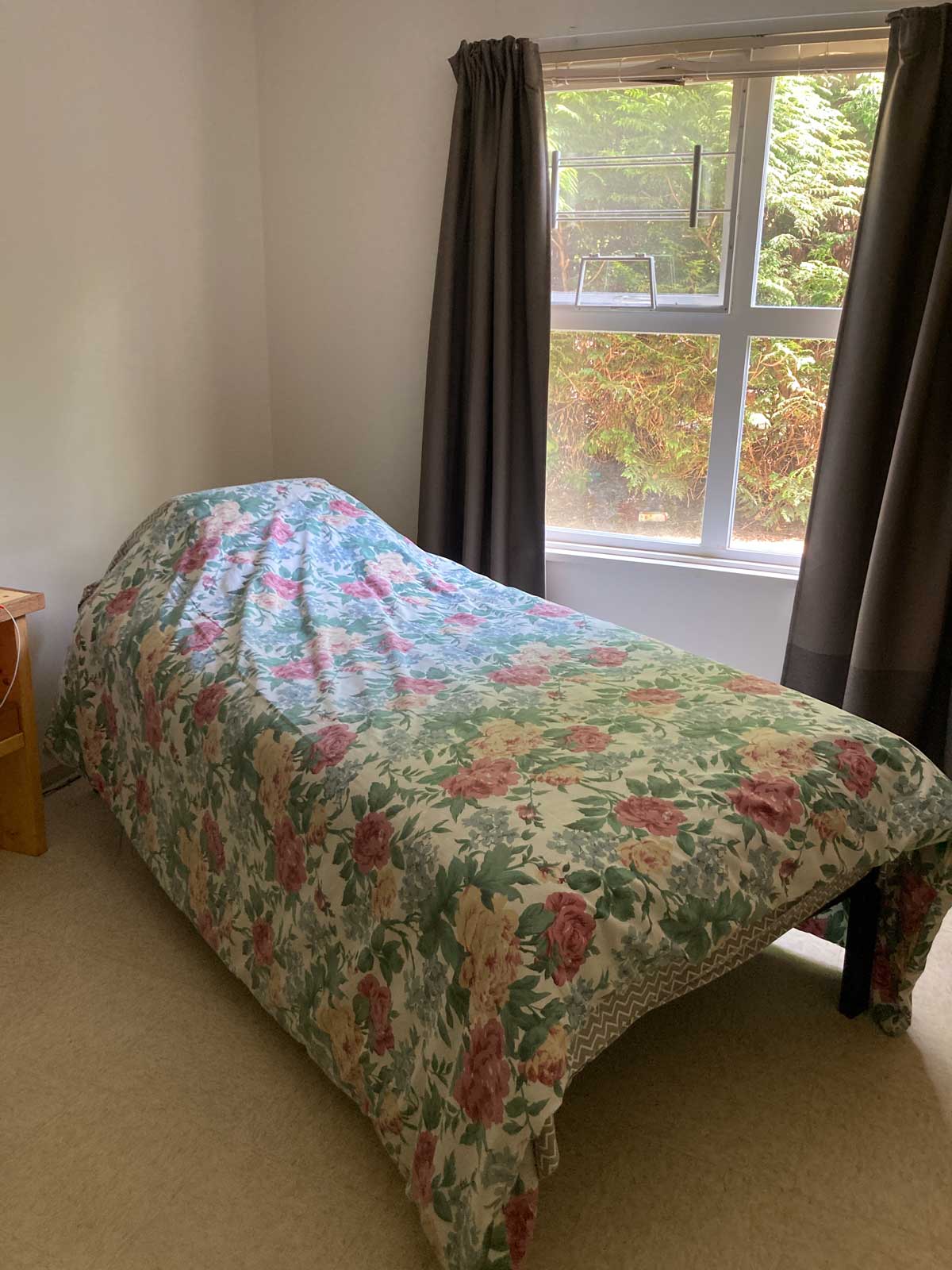 twin bed and pillow covered with floral duvet are dramatically angled into the corner of a small bedroom in senior housing. Open dark brown curtains reveal gold autumn leaves through the window.